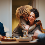 Two female friends at a dinner table looking happy, They are hugging. 